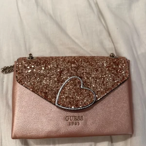 Rosa glittrig axelväska från Guess - Supersöt rosa axelväska från Guess med glittrigt lock och hjärtformad detalj framtill. Väskan har kedjerem i silver och flera fack inuti. 