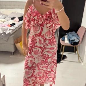 Blommig volangklänning med smala axelband - Säljer en superfin blommig klänning i vitt och rosa med volangdetaljer upptill och smala axelband. Klänningen har en luftig passform och ett härligt somrigt mönster, perfekt för varma dagar. Längden är mitt på vaden och tyget känns lätt och bekvämt.