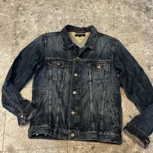 Mörkblå jeansjacka från Tommy Hilfiger - Snygg mörkblå jeansjacka från Tommy Hilfiger med klassisk passform. Jackan har två bröstfickor med knappar, sidofickor och silverfärgade knappar framtill. Tidlös design med diskret logga på bröstfickan och kontrastsömmar. Stl s