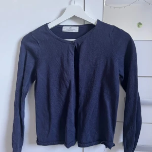 Mörkblå kofta från H&M - Snygg mörkblå kofta från H&M i storlek 134/140. Koftan har rund hals, långa ärmar och knappar framtill. Materialet är mjukt och stickat, perfekt för lager-på-lager. Enkel och stilren design som funkar till många olika outfits.