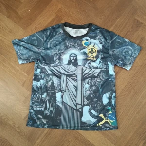 Brasil fotbollströja - Unik fotbollströja från Brasil med stort grafiskt tryck av Kristusstatyn och detaljerade mönster i blå och grå toner. Kortärmad modell med rund hals och Brasil-logga på bröstet. Materialet är lätt och andas, perfekt för match eller träning.