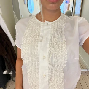 Vit kortärmad blus med spetsdetaljer - Supersöt vit blus med korta ärmar och dekorativa spetsdetaljer framtill. Blusen har pärlknappar och en rak passform, perfekt till jeans eller kjol. Materialet känns lätt och luftigt, och spetsen ger en romantisk vibe.