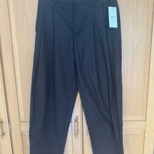 Mörk bruna wide tapered kostymbyxor - Mörkbruna kostymbyxor med wide fit och tapered ben, tillverkade i en återvunnen polyesterblandning. Byxorna har hällor för bälte, snygga fickdetaljer bak och en stilren look som passar till många olika outfits. Perfekt för dig som gillar trendiga och bekväma byxor. Helt oanvända! Rollas av innan de skickas!