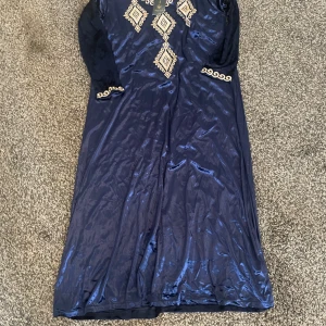 Marinblå långklänning Royal Mode. Arabiska kläder - Snygg marinblå långklänning från Royal Mode i glansigt material med guldfärgade broderade detaljer och mönster framtill samt vid ärmslut. Klänningen har långärmade ärmar och en elegant, rak siluett som ger en lyxig vibe. Det är mindre i storleken. Det står storlek XXL, vilket motsvarar storlek XL-L. 