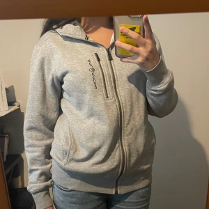 Grå hoodie sailracing hoodie med huva - Säljer en grå hoodie med dragkedja framtill och en extra bröstficka med zip. Perfekt på vintern då den är väldigt varm. Har storlek S i killmodell så den är lite oversized för kvinnor.