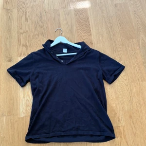 Svart t-shirt med krage från Lindex 170 - Svart t-shirt från Lindex med korta ärmar och cool krage med två knappar. T-shirten är i mjuk bomull och har en enkel, stilren look som funkar till det mesta. Passformen är normal och materialet känns skönt mot huden. Strl 170cm. Köpt för 299kr mitt pris 130kr.