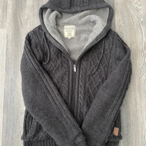 Mörkgrå stickad hoodie Portonova - Mysig mörkgrå stickad hoodie från Portonova med dragkedja och huva. Insidan är fodrad med grå fleece för extra värme. Perfekt för kyliga dagar när du vill vara både varm och stilren.