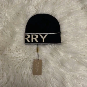 Svart Burberry mössa  - Snygg svart mössa från Burberry med stor vit logotyp runt kanten. Klassisk form och ribbad kant, perfekt för dig som vill ha en stilren accessoar med lyxig känsla. Materialet är stickat och mjukt, passar till kalla dagar.