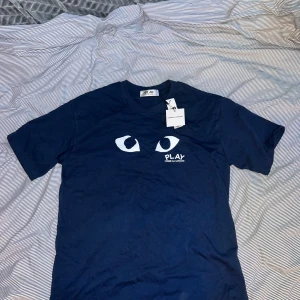 Blå Comme des Garçons Play t-shirt - Snygg marinblå t-shirt från Comme des Garçons Play med ikoniska vita ögon och logga på bröstet. Klassisk rund hals och korta ärmar. Tillverkad i 100% bomull för en skön och avslappnad känsla. Perfekt för dig som gillar streetwear med en twist. 0bs lnte 0rglnal!!