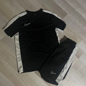 Svart Nike Dri-Fit fotbolls set  - Nike Dri-Fit fotbollströja i svart med vita paneler längs sidorna och axlarna. Kortärmad modell med rund hals och klassisk Nike-logga på bröstet. Tillverkad i lätt, ventilerande polyester som håller dig torr under träningen. Säljes tillsammans med matchande shorts. Man får med båda i priset! Ej hugget i sten
