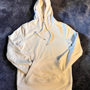 Beige hoodie med textdetalj - Säljer en stilren beige hoodie med broderad text på bröstet. Tröjan har huva med snörning, stor magficka och ribbade muddar vid ärmslut och nederkant. Perfekt för chill dagar och enkel att matcha med jeans eller joggers.