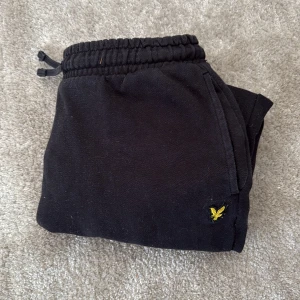 Svarta mjukisbyxor Lyle & Scott Junior - Svarta mjukisbyxor från Lyle & Scott Junior med elastisk midja och snörning. Byxorna har en broderad gul logga på ena benet och är tillverkade i mjukt bomullsmaterial. Perfekta för chill dagar eller när du vill vara bekväm.