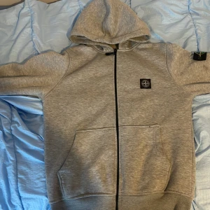 Grå hoodie från Stone Island - Säljer en grå hoodie från Stone Island med klassisk logopatch på ärmen och bröstet. Tröjan har huva, dragkedja framtill och två stora fickor. Mjuk bomullskvalitet och ribbade muddar. Perfekt för chill dagar och streetwear-stil.