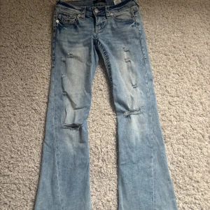 True Religion Joey Low Rise Flare Jeans - Snygga ljusblå jeans från True Religion, modell Joey Low Rise Flare. De har låg midja, utsvängda ben och slitna detaljer framtill för en cool vintagekänsla. Klassiska bakfickor med lock och silverknappar. Perfekta för dig som gillar retrostil! Använda cirka 3 ggr.
