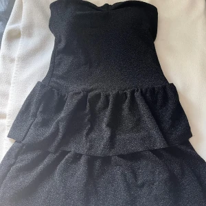 Svart glittrig kort klänning från Missay - Supersnygg svart glittrig klänning från Missay med volangdetalj i midjan. Axelbandslös design som ger en cool och festlig vibe. Perfekt för dig som vill sticka ut med lite extra glitter. Klänningen är kort och har en figurnära passform.perfekt för nyår! Helt oanvänd då lappen är kvar. Orginalpris ligger på 400kr