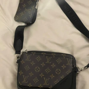  axelväska monogram brun - Säljer en snygg axelväska från  med klassiskt monogrammönster i brunt och beige. Väskan har bred svart axelrem med logga och en tillhörande liten plånbok med dragkedja. Perfekt för dig som vill ha en trendig och praktisk väska.
