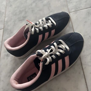 Adidas gazelle sneakers med rosa detaljer - Snygga Adidas sneakers i mörkblå mocka med klassiska rosa ränder och vit sula. Skorna har vita snören och rosa innerfoder som ger en fräsch kontrast. Perfekta för dig som vill ha en sportig och trendig look.
