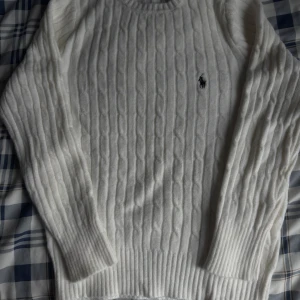 Vit kabelstickad tröja Polo Ralph Lauren - Vit kabelstickad tröja från Polo Ralph Lauren med klassisk rund hals och broderad logga på bröstet. Tröjan har långärmade ribbade muddar och är gjord i mjuk bomull. Perfekt för dig som gillar clean och tidlös stil. Storlek S använd 3 gånger, passar för dig som gillar slimfit 