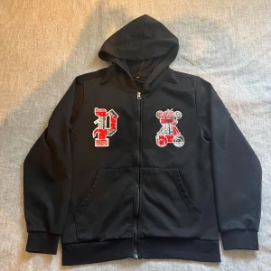 Svart hoodie - Svart hoodie med huva och hel dragkedja framtill. På bröstet finns ett stort rött och vitt P-tryck samt en nalle i samma färger. Perfekt för en chill och streetig look. Säljer för att jag ej är i behov av den längre. Vid snabb affär kan priset diskuteras!
