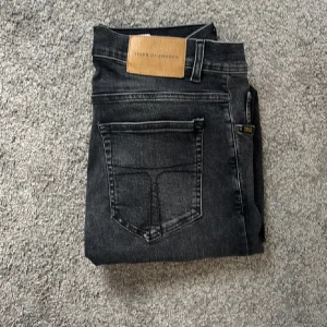 Svarta jeans från Tiger of Sweden - Snygga svarta jeans från Tiger of Sweden med klassisk femficksdesign och diskret tvättad look. Jeansen har normal passform, raka ben och är tillverkade i mjukt bomullsmaterial med lite stretch för extra komfort. Perfekt för dig som gillar stilrena och tidlösa jeans.
