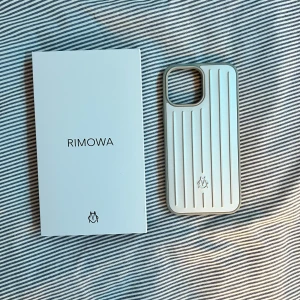 Rimowa telefonskal 13 PRO Max  - Säljer en stilren vit mobilväska från Rimowa med klassiska vertikala ränder och diskret logga framtill. Väskan har en hård, tålig yta och passar perfekt för dig som gillar minimalistisk design och vill ha något exklusivt till mobilen. iPhone 13 Pro Max