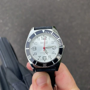 Regal svart-silver sportklocka - Snygg sportig klocka från Regal med svart gummiband och silverfärgad urtavla. Klockan har tydliga siffror, svart vridring och röd sekundvisare. Vattentät upp till 50 meter, perfekt för dig som gillar en stilren och funktionell accessoar.