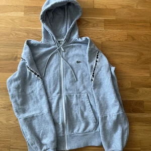 Grå hoodie från Lacoste med dragkedja - Snygg grå hoodie från Lacoste med dragkedja och huva. Klassisk passform, snörning i huvan och logoband på ärmarna. Mjuk insida och två stora fickor framtill. Perfekt för chill dagar eller när du vill ha en sportig look.