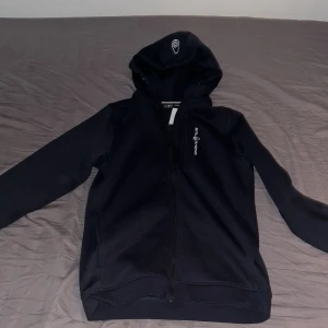 Svart Sail Racing hoodie med dragkedja - Svart hoodie från Sail Racing i storlek 176. Tröjan har hel dragkedja framtill, huva med logga och en bröstficka med dragkedja. Snyggt Sail Racing-tryck på bröstet. Perfekt för dig som gillar sportig och stilren look. Tröjan är dessutom helt oanvänd.