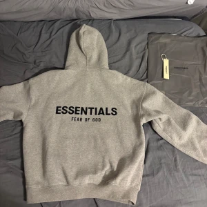 Grå Essentials Fear of God hoodie - Snygg grå hoodie från Essentials Fear of God med svart logga på bröstet och stor text på ryggen. Klassisk känguruficka, huva och relaxed fit. Perfekt för dig som gillar streetwear och vill ha en stilren look.