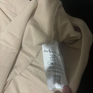 Beige t-shirt från Polo Ralph Lauren - Snygg beige t-shirt från Polo Ralph Lauren med klassisk rund hals och korta ärmar. T-shirten är i 100% bomull och har den ikoniska Polo-loggan broderad på bröstet. Perfekt för dig som gillar stilrena och enkla plagg med en touch av lyx.