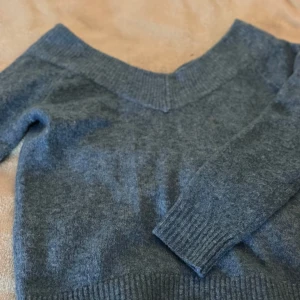 Grå stickad tröja med v-ringning H&M - Mysig blå stickad tröja från H&M i storlek S. Tröjan har en bred v-ringad hals och långa ärmar med ribbade muddar. Perfekt för kyliga dagar och enkel att matcha med jeans eller kjol. Materialet är mjukt och ger en skön känsla.