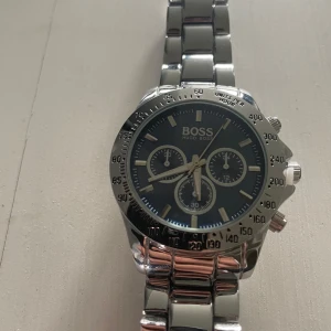 Silverfärgad Hugo Boss herrklocka - Stilren herrklocka  silverfärgat armband i metall och mörkblå urtavla. Klockan har tre mindre visartavlor, kronograf och tydliga markeringar runt boetten. Perfekt accessoar för dig som gillar klassisk design med modern touch.