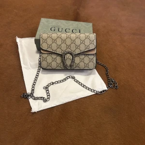 Gucci Dionysus beige axelväska - Säljer en Gucci Dionysus axelväska i beige canvas med klassiskt GG-mönster och snygg silverfärgad kedja. Väskan har ikoniskt spänne framtill och detaljer i skinn och mocka. Perfekt storlek för det viktigaste och ger en lyxig touch till din outfit.