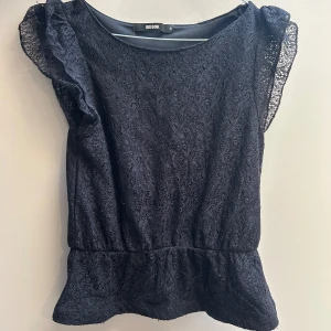 Svart spetsblus från Bik Bok - Supersöt svart blus från Bik Bok i spets med volangdetaljer på axlarna. Blusen har en feminin siluett med lätt markerad midja och är ärmlös. Perfekt att styla med jeans eller kjol för en trendig look.