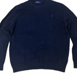 Mörkblå stickad tröja Polo Ralph Lauren - Snygg mörkblå stickad tröja från Polo Ralph Lauren med rund halsringning och diskret grön broderad logga på bröstet. Tröjan har ribbade muddar vid ärmslut och nederkant, perfekt för lager-på-lager-stil.