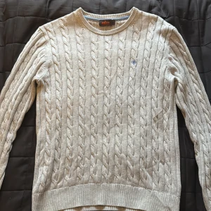 Grå kabelstickad tröja från Morris - Säljer en grå kabelstickad tröja från Morris i 100% pima bomull. Tröjan har rund hals, lång ärm och en liten blå broderad detalj på bröstet. Perfekt för dig som gillar klassisk och stilren look.