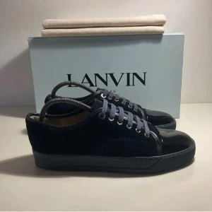 Mörkblå sneakers från Lanvin - Snygga mörkblå sneakers från Lanvin med ovandel i mocka och tå i blankt skinn. Skorna har rund tå, platt sula och snörning med matchande blå skosnören. Perfekta för dig som gillar stilrena och exklusiva sneakers med lyxig känsla.