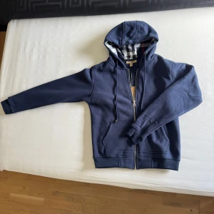 Marinblå hoodie från Burberry - Snygg marinblå hoodie från Burberry med dragkedja, huva med klassiskt rutigt foder och broderad logga på bröstet. Tröjan har känguruficka, snörning i huvan och är tillverkad i mjuk bomull. Perfekt för en chill och stilren look.