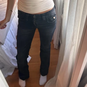 Svarta bootcut jeans med fickdetaljer - Säljer ett par svarta jeans med bootcut passform och snygga ficklock med knappar bak. Jeansen har låg midja och klassisk femficksdesign. Materialet är mjukt denim som sitter skönt och ger en avslappnad vibe.