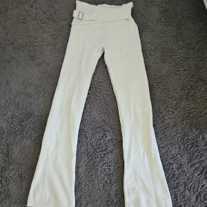 Vita stickade byxor med bälte XS - Snygga vita stickade byxor i storlek XS med låg midja och bred ribbad kant upptill. Byxorna har ett dekorativt bälte med metallspänne och raka ben med ribbade avslut. Perfekta för dig som gillar en chill och trendig look.