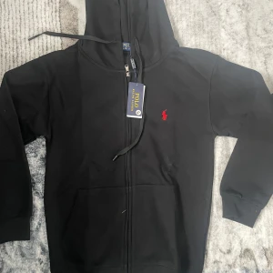 Svart hoodie från Polo Ralph Lauren - Svart hoodie från Polo Ralph Lauren med dragkedja, huva och snörning. Klassisk röd broderad logga på bröstet och känguruficka framtill. Tillverkad i mjuk bomull, perfekt för chill dagar eller när du vill ha en clean look.