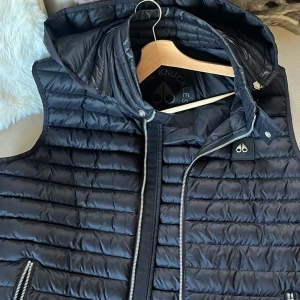 Svart dunväst från Moose Knuckles - Säljer en svart dunväst från Moose Knuckles med avtagbar huva och snygga silverfärgade dragkedjor. Säljer västen då jag inte använder den längre 