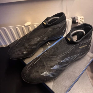 Adidas Predator svarta fotbollsskor - Svarta Adidas Predator fotbollsskor i storlek 42 med stilren design och diskreta detaljer. Skorna har en strumpliknande ovandel i syntetmaterial och mönstrad yta för extra grepp. Perfekta för dig som vill dominera på planen. Ett hål