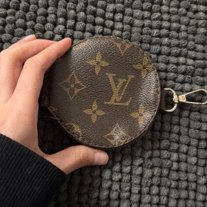 rund nyckel plånbok som kan sättas som nyckelring  - Säljer en rund plånbok som kan sättas som nyckel ring eller sättas på din Väsk kedja i brunt och guld. Plånboken har en praktisk dragkedja med logga på. Perfekt för enstaka kort, nycklar, AirPods eller småsaker. (Obs: den är lite rostig på dragkedjan se bild)