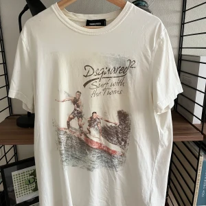 Vit Dsquared2 t-shirt med surftryck XL - Vit t-shirt från Dsquared2 i bomull med coolt surfmotiv och texten 'Shirt with the Twins' på bröstet. Klassisk passform, rund hals och korta ärmar. Perfekt för dig som gillar streetstyle och vill sticka ut med en unik printad tee.