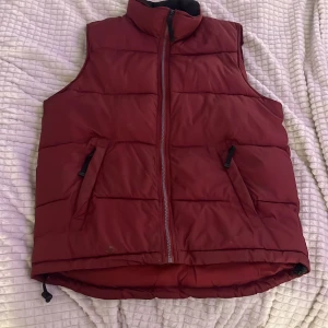 Vinröd dunväst från MyWear M - Snygg vinröd dunväst från MyWear i storlek M. Västen har hög krage, två fickor med dragkedja och quiltad design. Perfekt att slänga över en hoodie för extra värme. Justerbar nertill med snörning för bästa passform. Några fläckar framtill men borde gå bort i tvätt.