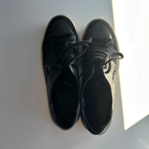 Svarta sneakers från Axel Arigato - Svarta sneakers i skinn från Axel Arigato med vit sula och klassisk låg profil. Skorna har snörning och en broderad fågel på sidan. Perfekta för en clean och stilren look.