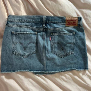 Levi's jeanskjol med rå kant, strl 28 - Snygg blå jeanskjol från Levi's med klassiska bakfickor, slitning framtill och rå, fransig kant nertill. Kjolen har bälteshällor och knappstängning. Perfekt för dig som gillar en avslappnad och trendig stil.