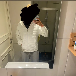 Vit stilren cardigan/hybridjacka - Säljer en stilren vit pufferjacka med huva och dragkedja framtill. Jackan har quiltade partier på kroppen och släta ärmar, samt två fickor med dragkedja. Perfekt för dig som vill ha en clean och modern look. Diskuterar gärna pris!