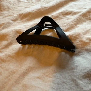 Svart hårklämma i matt plast - Säljer en stilren svart hårklämma med matt finish och robust fjäder. Klämman har en trekantig form och breda grepp som håller håret på plats hela dagen. Perfekt för att snabbt fixa frisyren och ge en clean look.